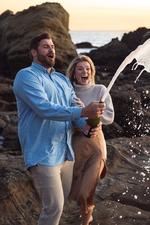 Champagne sprays as a couple laugh on the coastal rocks