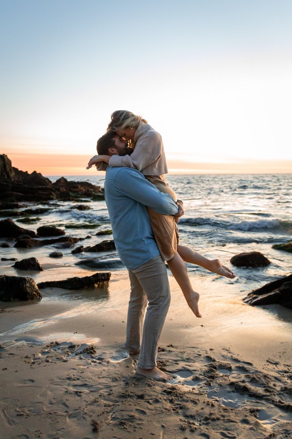 A man lifts his partner off her feet on the beach at sunset