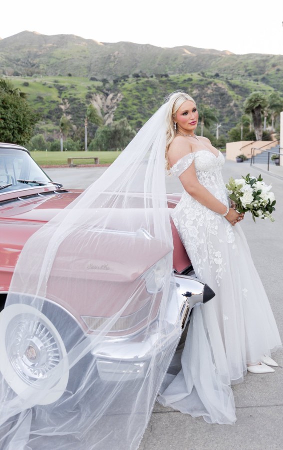 A bride in a long veil beside a vintage car, hills behind her