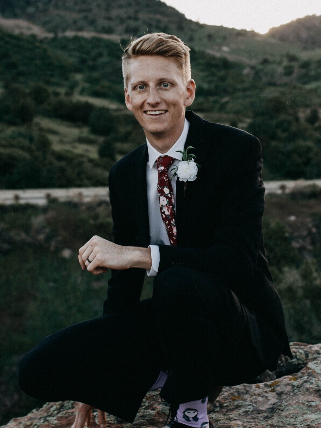 Daniel Lindeen crouched on a rock in a dark suit, hills behind him